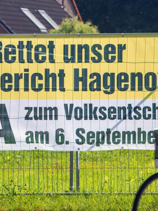 Plakat hängt an einem Zaun, eine Frau auf einem Rad fährt vorbei, Aufschrift auf dem Plakat: "Rettet unser Amtsgericht Hagenow! Ja zum Volksentscheid am 6. September"