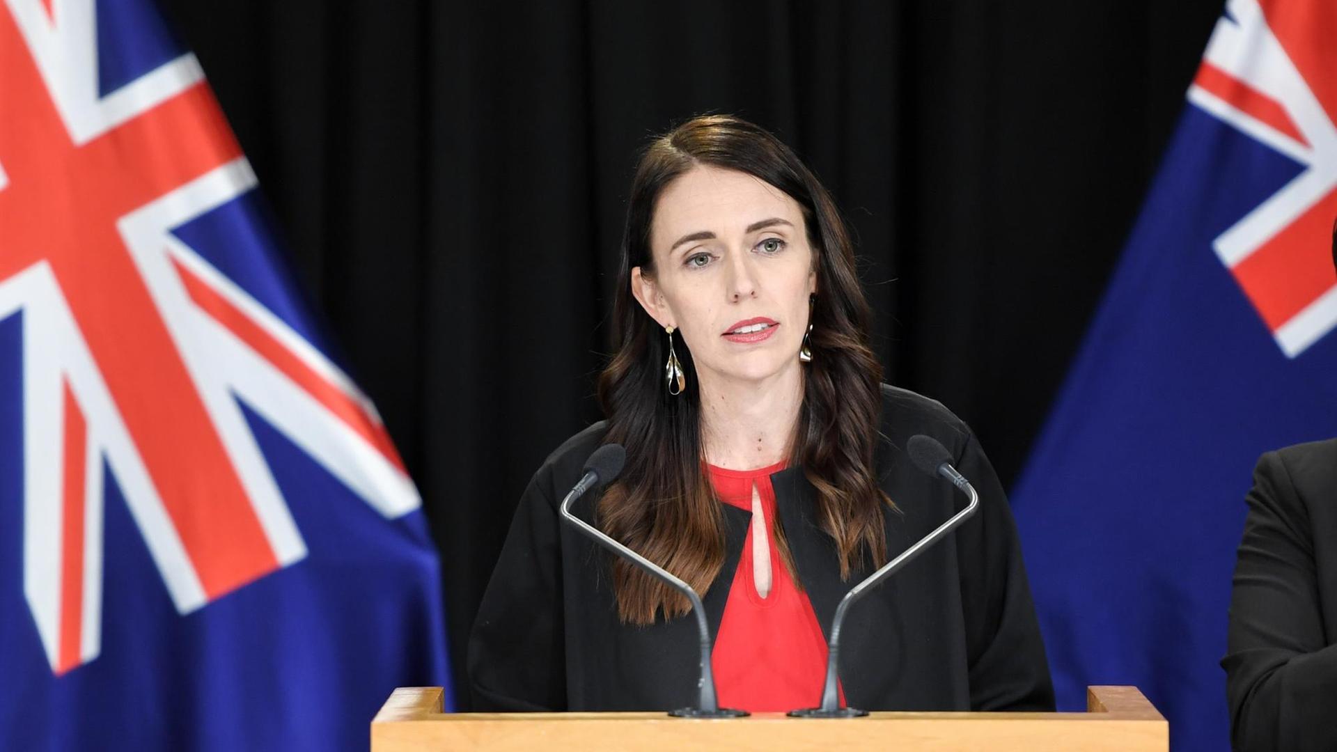 Porträt der neuseeländischen Premierministerin Jacinda Ardern am Rednerpult, umsäumt von Flaggen. Porträt der neuseeländischen Premierministerin Jacinda Ardern am Rednerpult, umsäumt von Flaggen.