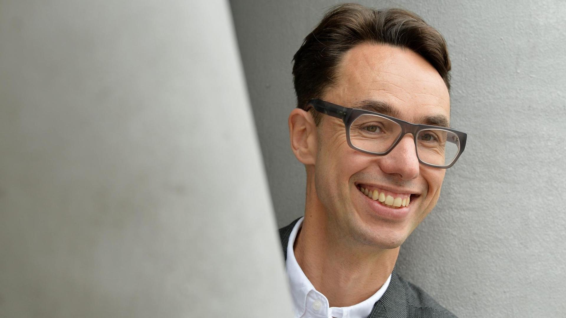 Hinter einem grauen Pfeiler schaut ein Mann mit dunklen zurückgekämmten Haaren und großer Brille und einem freundlichen Gesicht hervor. Er trägt ein weißes Hemd und eine Anzugjacke. Hinter einem grauen Pfeiler schaut ein Mann mit dunklen zurückgekämmten Haaren und großer Brille und einem freundlichen Gesicht hervor. Er trägt ein weißes Hemd und eine Anzugjacke.