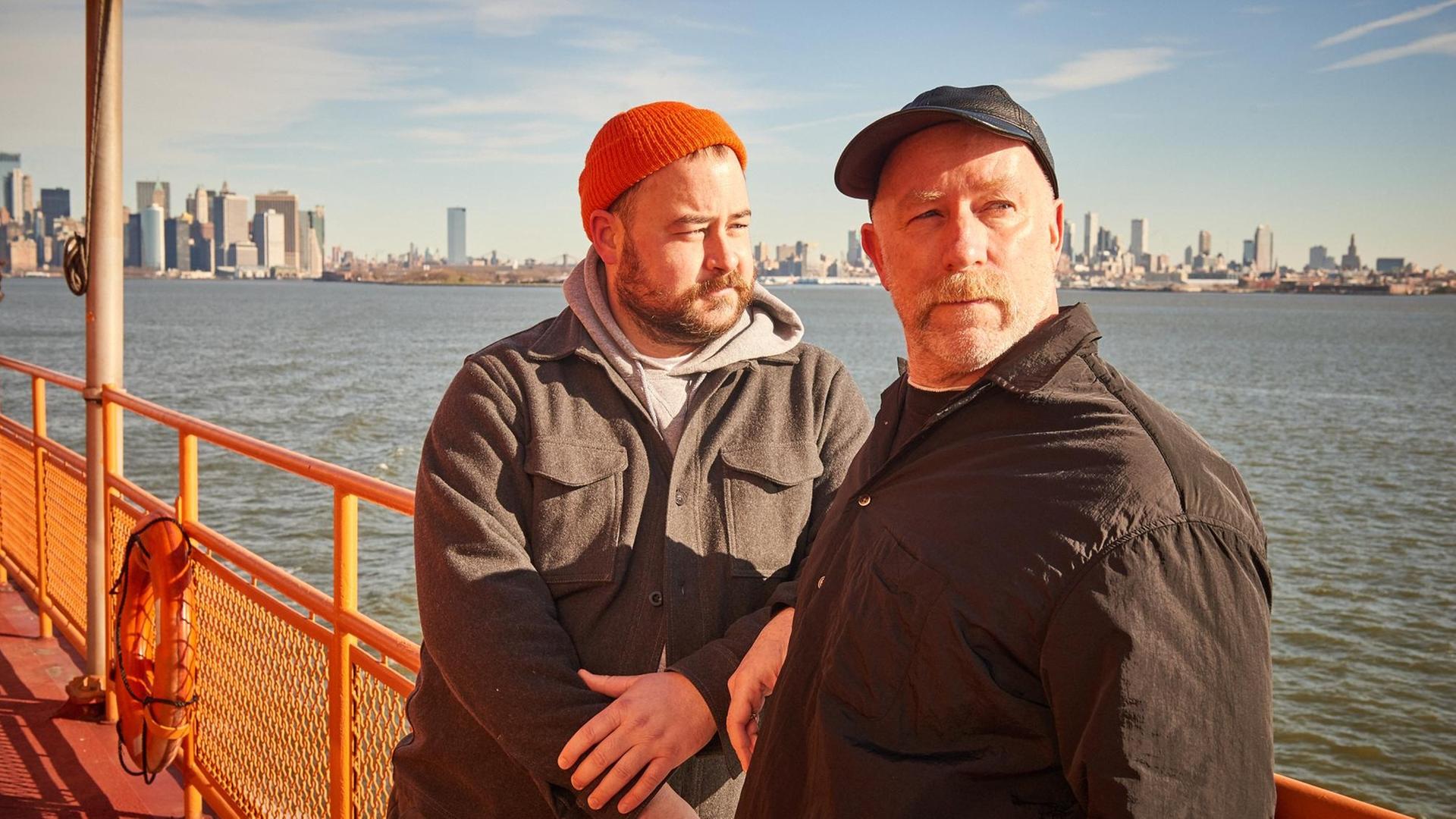 An Bord einer Fähre stehen zwei Männer mit Mützen und warmen Jacken an der Reling. Im Hintergrund sind die Hochhäuser von New York City zu sehen. An Bord einer Fähre stehen zwei Männer mit Mützen und warmen Jacken an der Reling. Im Hintergrund sind die Hochhäuser von New York City zu sehen.