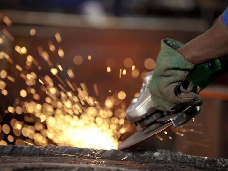 Symbolbild: Ein Afrikaner schleift einen Industriemagneten, zu sehen ist ein Stück vom Arm, die Hand in einem Handschuh, die Schleifmaschine, die Funken schlägt, und ein Stück vom Magneten.