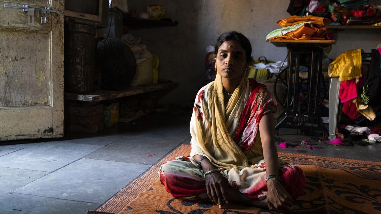Eine junge Frau im Sari mit traurigem Gesicht sitzt im Schneidersitz in ihrem Wohnhaus auf einem Teppich.