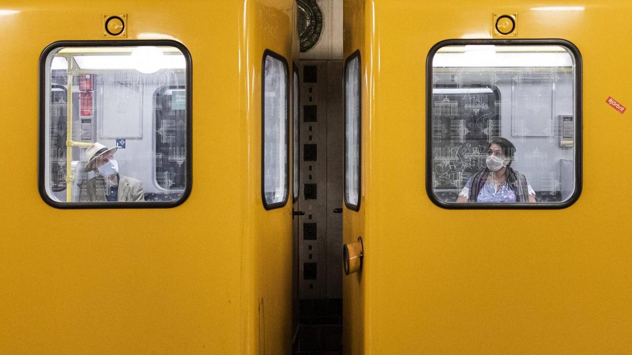 Zwei Personen mit Mund-Nasen-Maske sitzen in unterschiedlichen Waggons der Berliner U-Bahn. Zwei Personen mit Mund-Nasen-Maske sitzen in unterschiedlichen Waggons der Berliner U-Bahn.