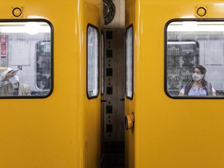 Zwei Personen mit Mund-Nasen-Maske sitzen in unterschiedlichen Waggons der Berliner U-Bahn.