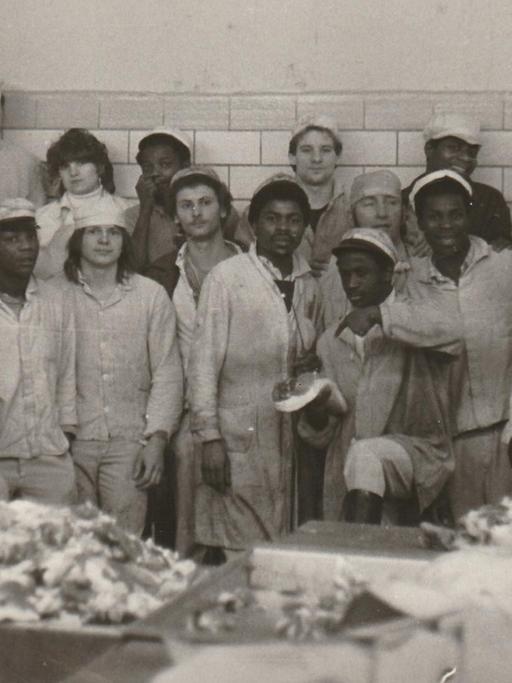 Gruppenfoto mit DDR-Vertragsarbeitern aus Mosambik im VEB Fleischkombinat Berlin 1982. Männer und Frauen in Arbeitskleidung posieren für ein Gruppenbild in einem fleischverarbeitenden Betrieb.