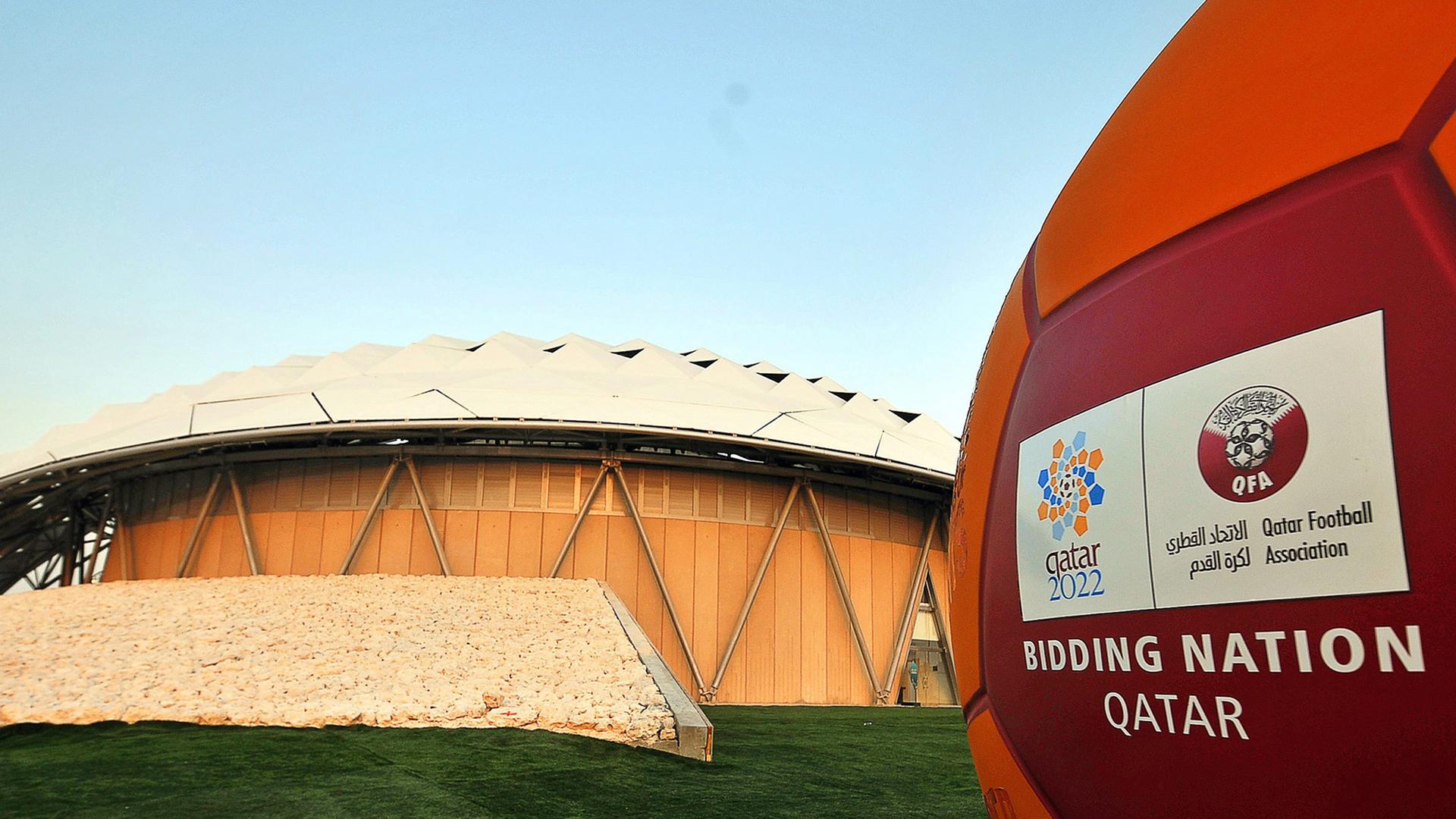 Ein roter Ball mit dem Logo Katars als Bewerber für die Austragung der Fußball-WM 2022, im Hintergrund ein Fußballstadion