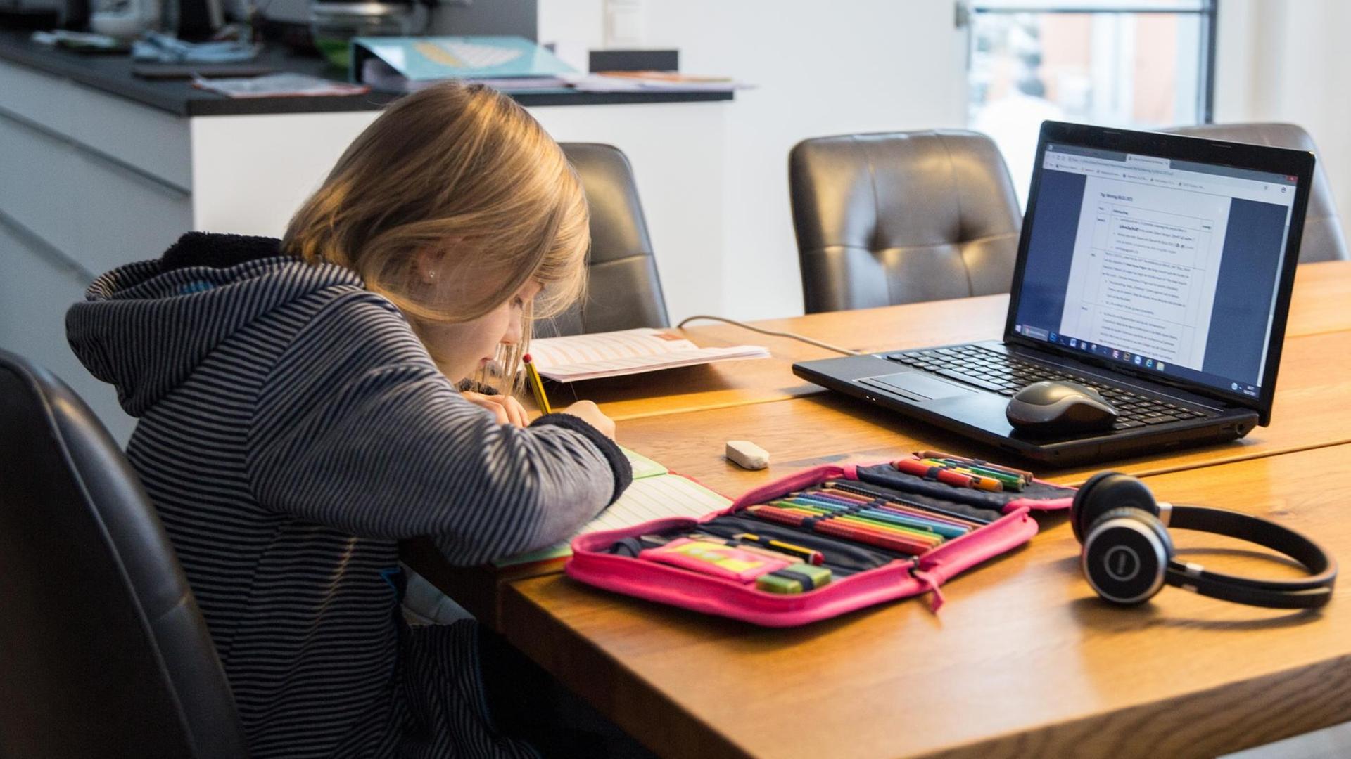 Ein Kind sitzt an einem Küchentisch und löst Aufgaben in einem Schulheft. Auf dem Tisch steht ein aufgeklappter Laptop, neben dem Rechner liegen Bücher und Hefte mit weiteren Schulutensilien für das Homeschooling.