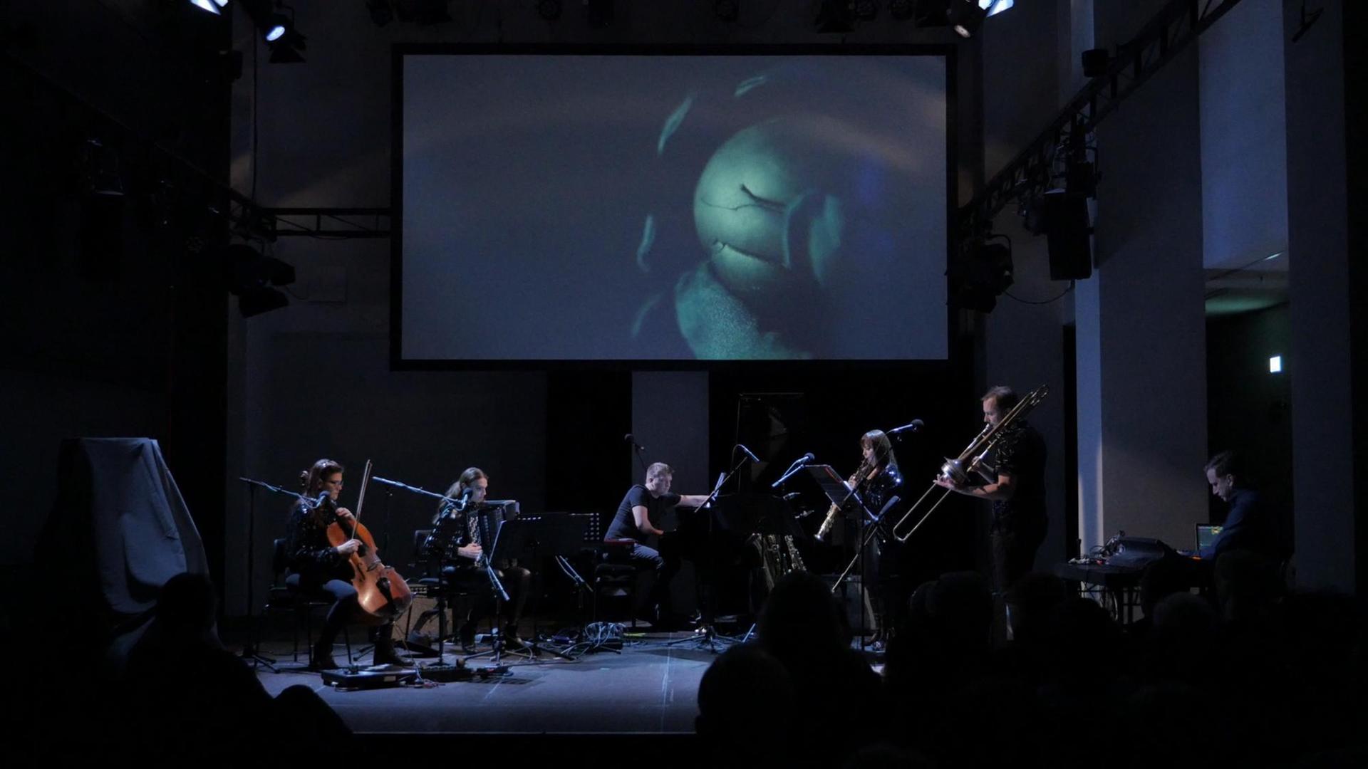 Sechs Musikerinnen und Musiker performen auf einer dunkelblau ausgeleuchteten Bühne. Über Ihnen im hängt eine Leinwand mit einer Projektion.