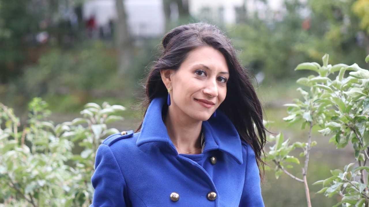 Eine schmale Frau mit dunklen langen Haaren in einem indigo-blauen Mantel. Der Kragen ist hochgeklappt. Sie steht in einem Park. Eine schmale Frau mit dunklen langen Haaren in einem indigo-blauen Mantel. Der Kragen ist hochgeklappt. Sie steht in einem Park.