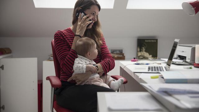 Eine Frau sitzt telefonierend am Schreibtisch, auf dem Schoß ihr Baby