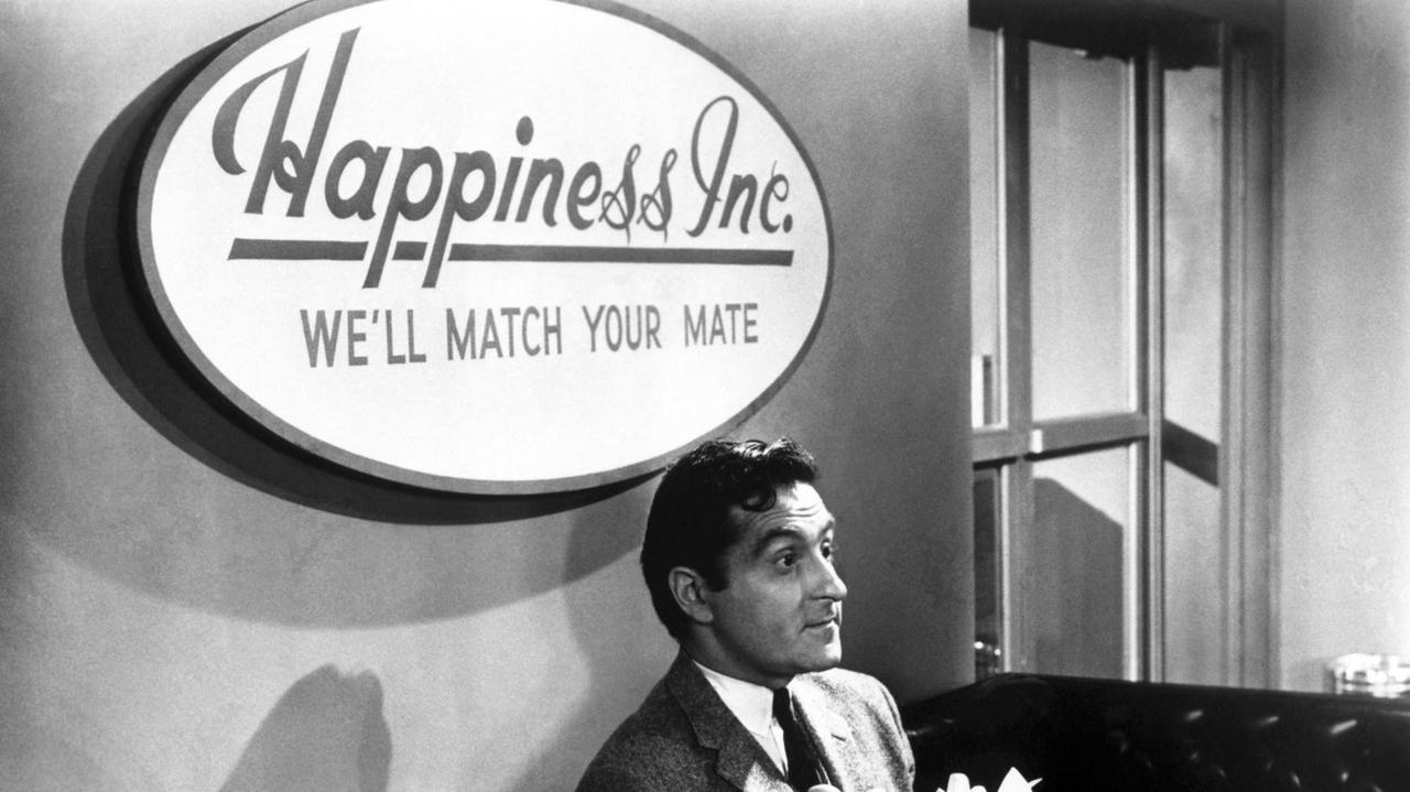 Szene aus der Kömödie "For Singles only" - ein gutaussehender Mann sitzt auf einem Sofa unter einem Schild an der Wand: "Happiness Inc. - We'll find your mate"