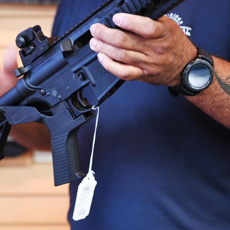 Das Bild zeigt den Oberkörper des Verkäufers mit tätowiertem Arm, der das Gewehr in den Händen hält. Er trägt zudem ein Halfter mit Pistole am Gürtel.