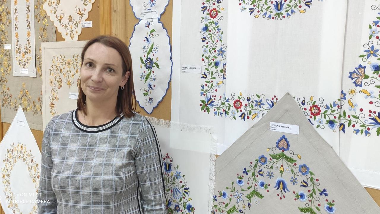 Kamila Soroko, die Kulturhausleiterin, steht im grau karierten Pullover vor einer Wand, an der unterschiedliche traditionelle Stickereien mit Blumenmustern aufgehängt sind. Kamila Soroko, die Kulturhausleiterin, steht im grau karierten Pullover vor einer Wand, an der unterschiedliche traditionelle Stickereien mit Blumenmustern aufgehängt sind.