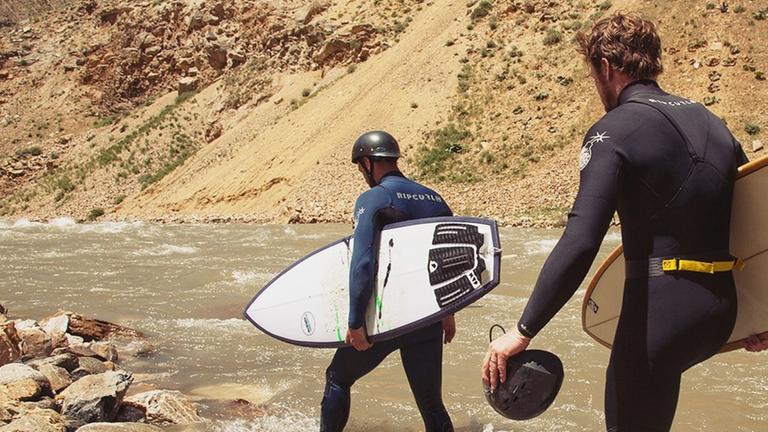 Zwei Männer in Neoprenanzügen und mit Surfboards in den Händen waten durch einen Fluss in Afghanistan. Zwei Männer in Neoprenanzügen und mit Surfboards in den Händen waten durch einen Fluss in Afghanistan.