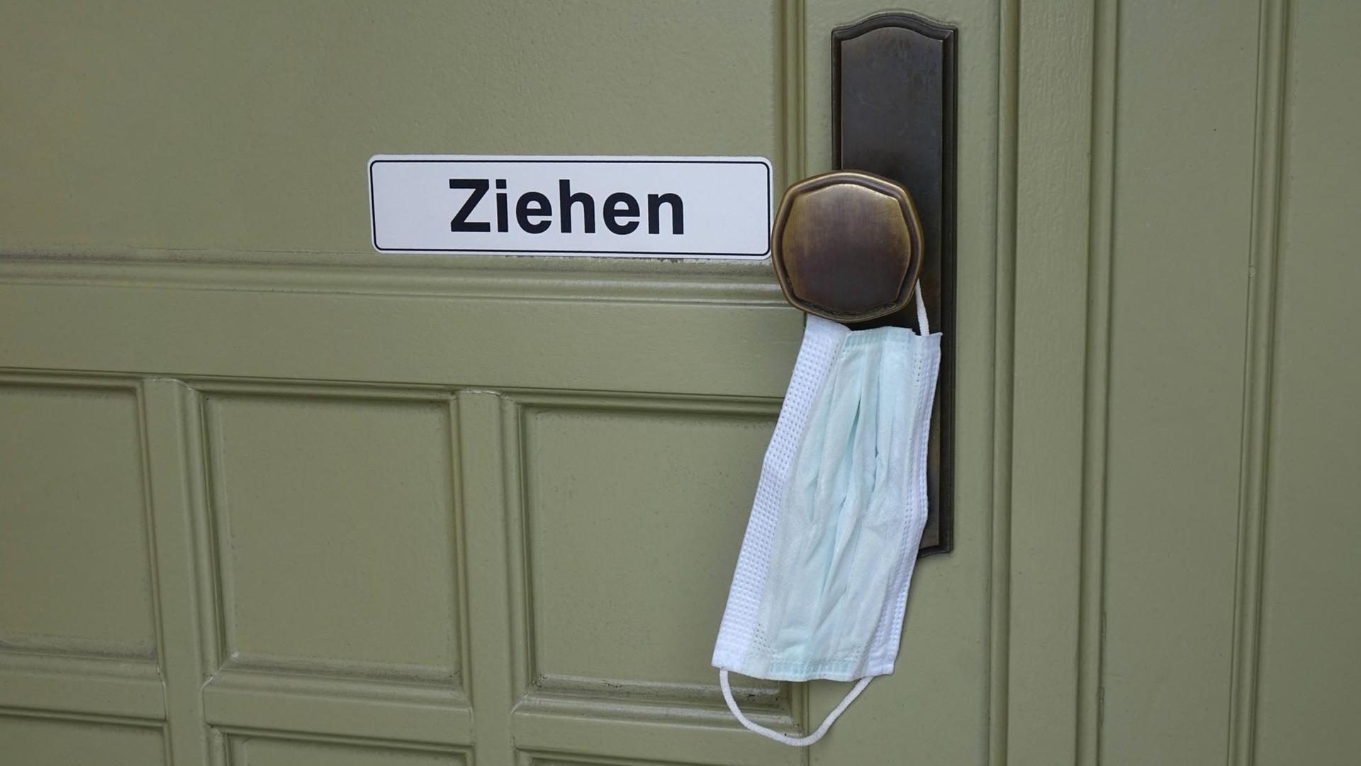 An einer Türklinke hängt zum Öffnen eine Atemschutzmaske An einer Türklinke hängt zum Öffnen eine Atemschutzmaske