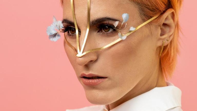 Eine Frau mit pfirsichfarbenem kurzen Haar trägt ein feines goldenes Gestell über Stirn und Nase. Hellblaue Blumen wirken wie Wimpernverlängerungen. Sie trägt eine weiße Bluse mit Kragen, steht vor rosafarbenem Hintergrund und blickt mit konzentriertem Blick seitlich an der Kamera vorbei. Eine Frau mit pfirsichfarbenem kurzen Haar trägt ein feines goldenes Gestell über Stirn und Nase. Hellblaue Blumen wirken wie Wimpernverlängerungen. Sie trägt eine weiße Bluse mit Kragen, steht vor rosafarbenem Hintergrund und blickt mit konzentriertem Blick seitlich an der Kamera vorbei.