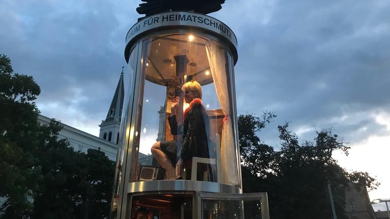 Eine Litfaßsäule aus Glas, in ihr sitze eine blonde Frau. Eine Litfaßsäule aus Glas, in ihr sitze eine blonde Frau.