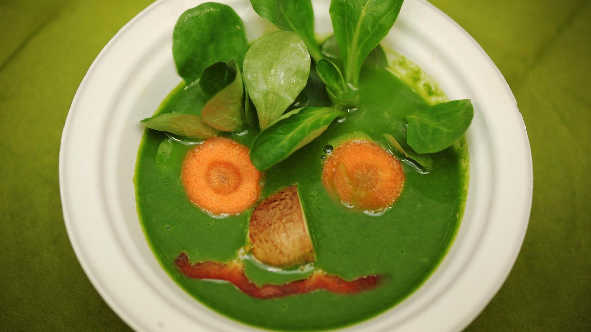 Eine vegane Spinat-Suppe, auf die aus Gemüse wie Spinatblätter und Möhren ein Gesicht gezeichnet wurde. Eine vegane Spinat-Suppe, auf die aus Gemüse wie Spinatblätter und Möhren ein Gesicht gezeichnet wurde.