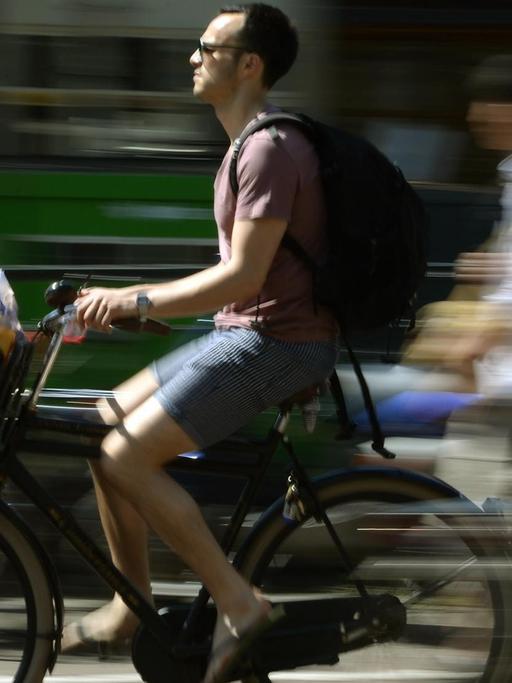 Ein Mann transport in seinem Fahrradkorb vorne am Lenker seine Einkäufe, während entlang einer der Amsterdamer Grachten fährt.