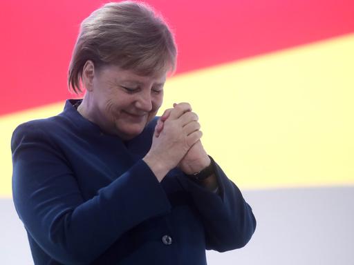 Angela Merkel hält die Hände gefaltet in die Höhe, hat den Kopf geneigt und die Augen kurz geschlossen.