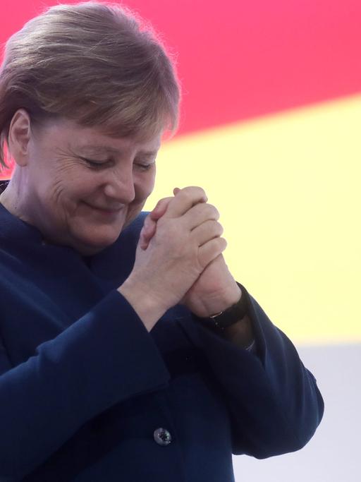 Angela Merkel hält die Hände gefaltet in die Höhe, hat den Kopf geneigt und die Augen kurz geschlossen.