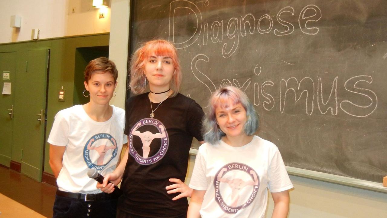 Lisa Wernicke, Fredericke Sonntag und Marie Römer von der Aktivistinnen-Gruppe "Medical Students for Choice" stehen vor einer Tafel, auf der "Diagnose Sexismus" geschrieben steht.