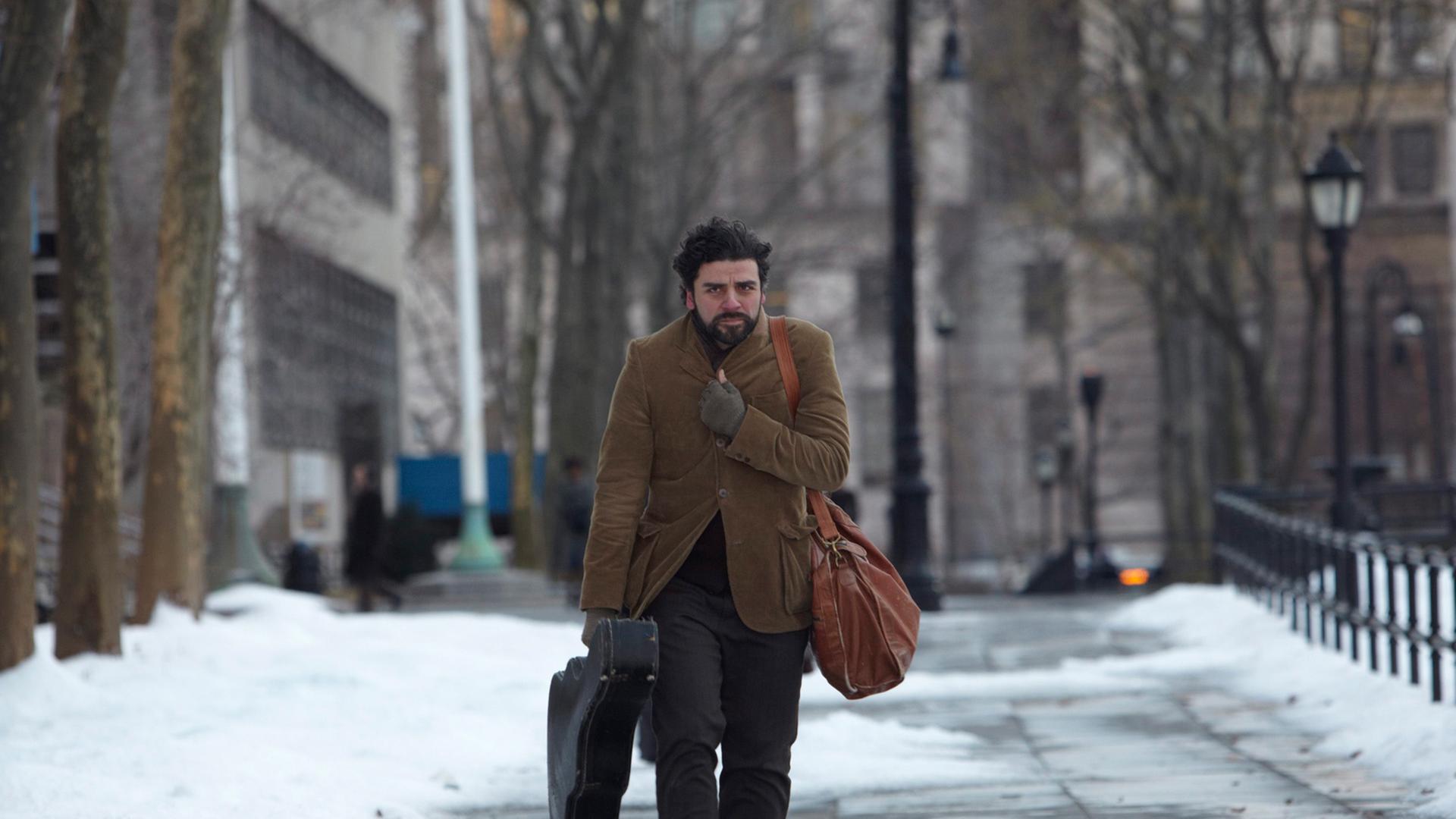 Film-Still: US-Schauspieler Oscar Isaac spaziert in "Inside Llewyn Davis" durch eine schneebedeckte Straße, Gitarre in der Hand, sich mit dem Sakko gegen die Kälte schützend Film-Still: US-Schauspieler Oscar Isaac spaziert in "Inside Llewyn Davis" durch eine schneebedeckte Straße, Gitarre in der Hand, sich mit dem Sakko gegen die Kälte schützend