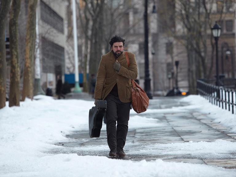 Film-Still: US-Schauspieler Oscar Isaac spaziert in "Inside Llewyn Davis" durch eine schneebedeckte Straße, Gitarre in der Hand, sich mit dem Sakko gegen die Kälte schützend