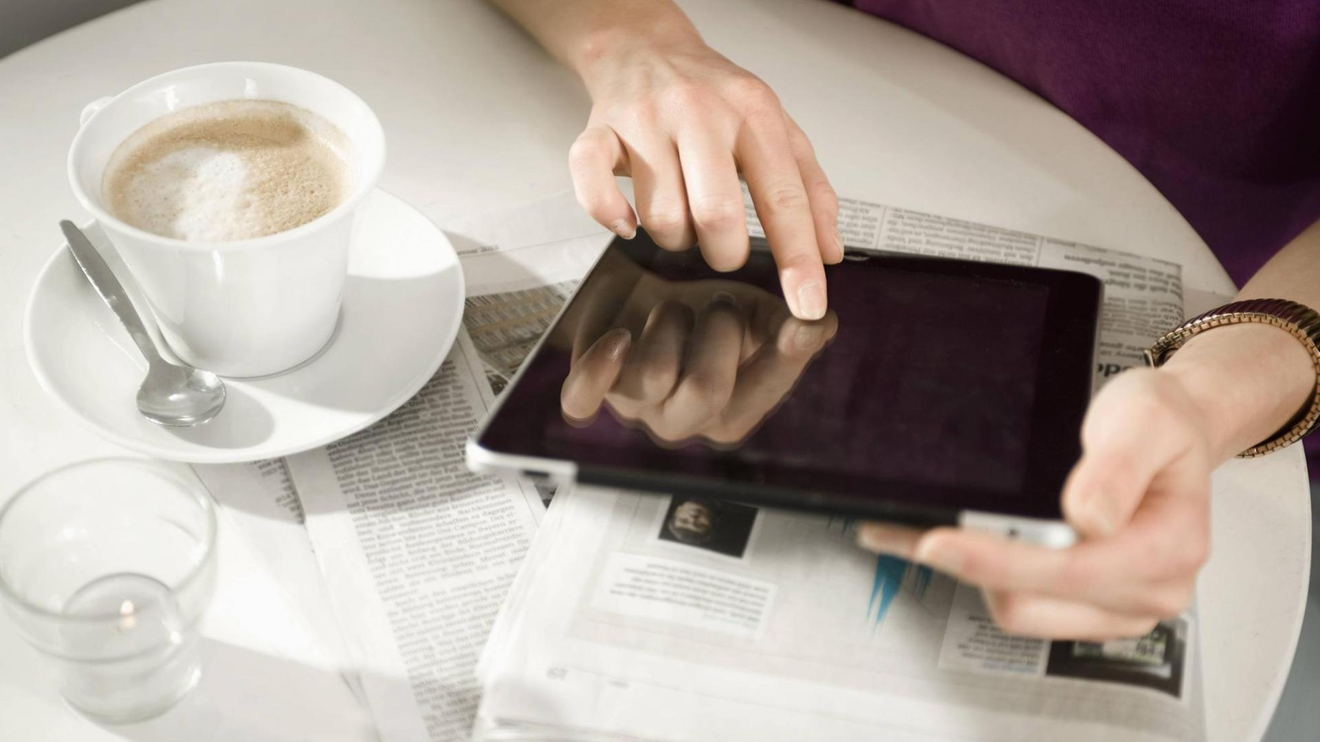Eine Frau sitzt in einem Café und benutzt ein Tablet, das auf auf einer Zeitung liegt. Eine Frau sitzt in einem Café und benutzt ein Tablet, das auf auf einer Zeitung liegt.