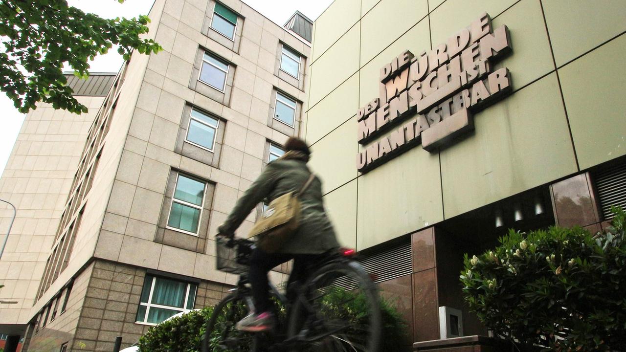 Eine Person fährt auf dem Fahrrad an der Fassade der Staatsanwaltschaft im Justizzentrum Frankfurt mit der Skulptur vorbei. Eine Person fährt auf dem Fahrrad an der Fassade der Staatsanwaltschaft im Justizzentrum Frankfurt mit der Skulptur vorbei.