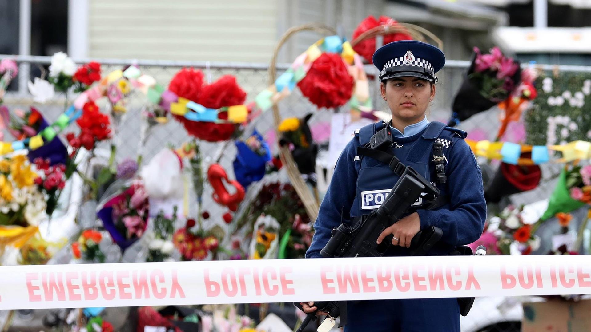 Eine Polizistin steht vor dem abgesperrten Tatort des Terroranschlags in Christchurch, hinter ihr sind zahlreiche niedergelegte Blumen zu sehen. Eine Polizistin steht vor dem abgesperrten Tatort des Terroranschlags in Christchurch, hinter ihr sind zahlreiche niedergelegte Blumen zu sehen.