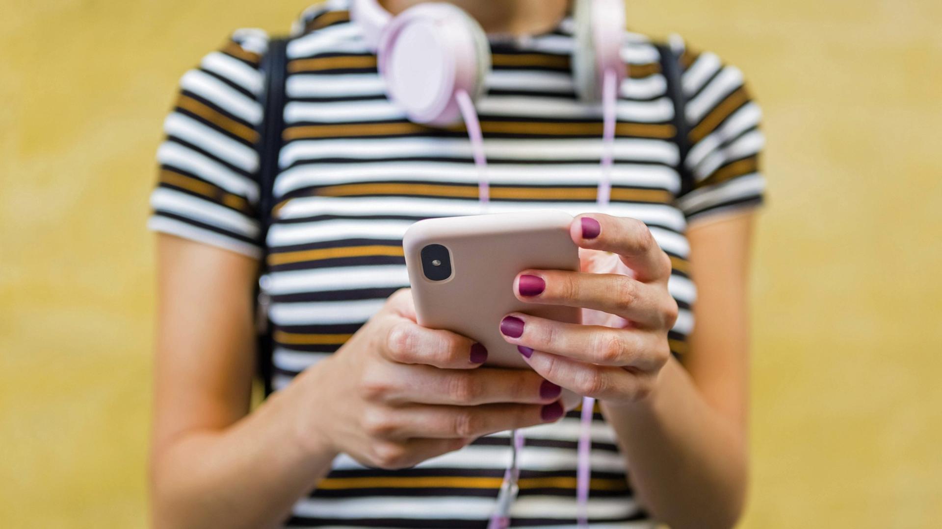 Eine Frau mit gestreiftem T-Shirt hält ein Smartphone in den Händen, um den Hals trägt sie Kopfhörer. Eine Frau mit gestreiftem T-Shirt hält ein Smartphone in den Händen, um den Hals trägt sie Kopfhörer.