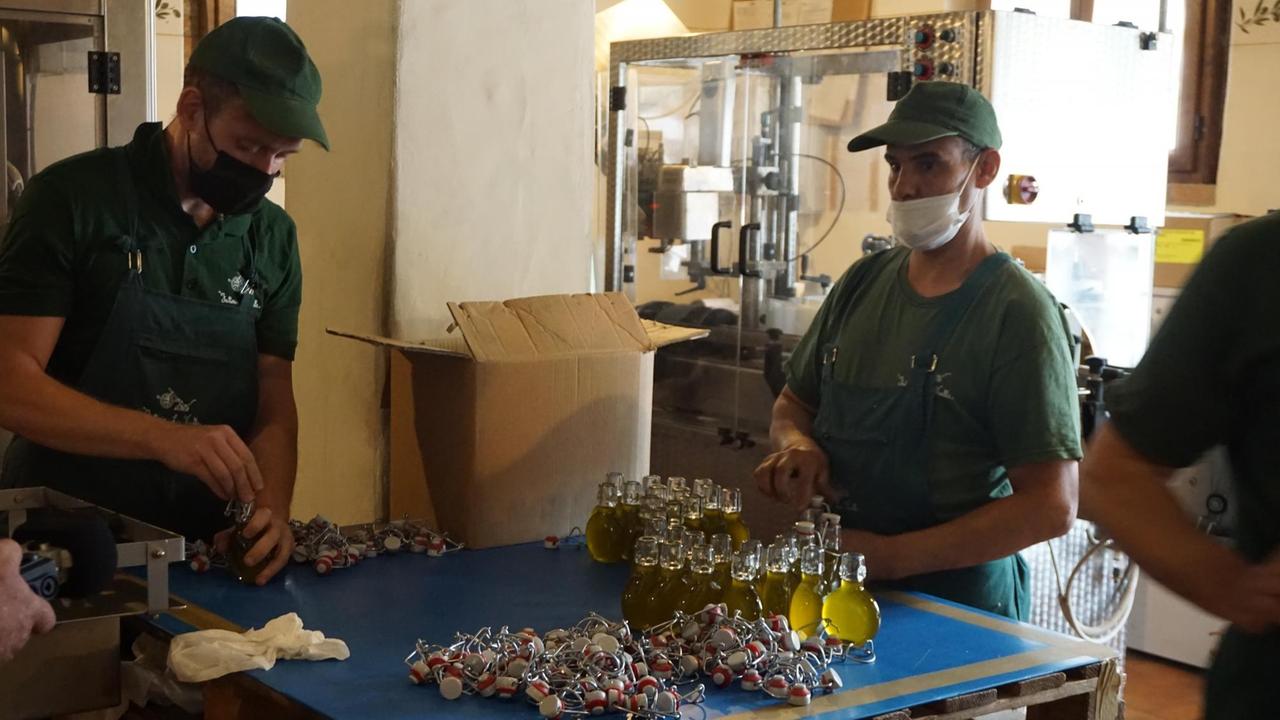 Arbeiter mit grünen Käppis und gleichfarbigen T-Shirts setzen Schnappverschlüsse auf mit Olivenöl gefüllte kleine Flaschen.