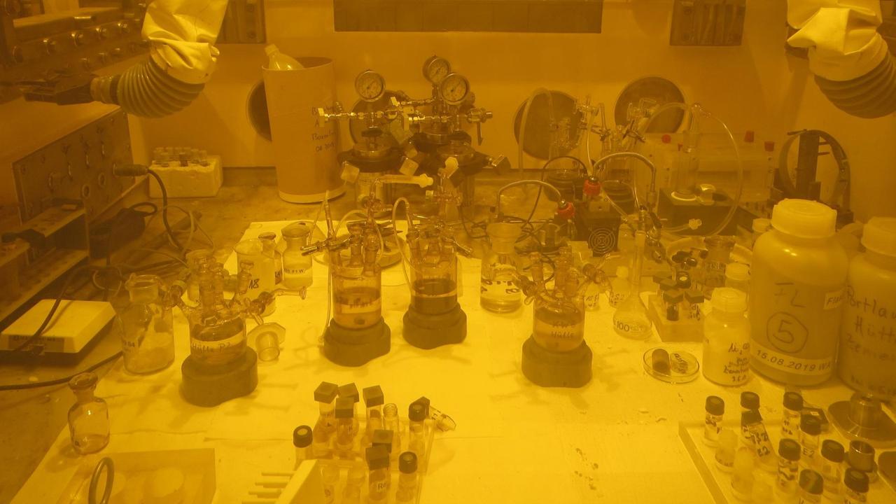Hinter einer dicken Bleiglasscheibe sieht man einen chemischen Versuchsaufbau mit Glaskolben, Schläuchen und Behältern mit Proben und Reagenzien, oben die Griffenden den Manipulatoren. Das Bild ist durch den Effekt der Bleiglasscheibe gelb eingefärbt. Hinter einer dicken Bleiglasscheibe sieht man einen chemischen Versuchsaufbau mit Glaskolben, Schläuchen und Behältern mit Proben und Reagenzien, oben die Griffenden den Manipulatoren. Das Bild ist durch den Effekt der Bleiglasscheibe gelb eingefärbt.
