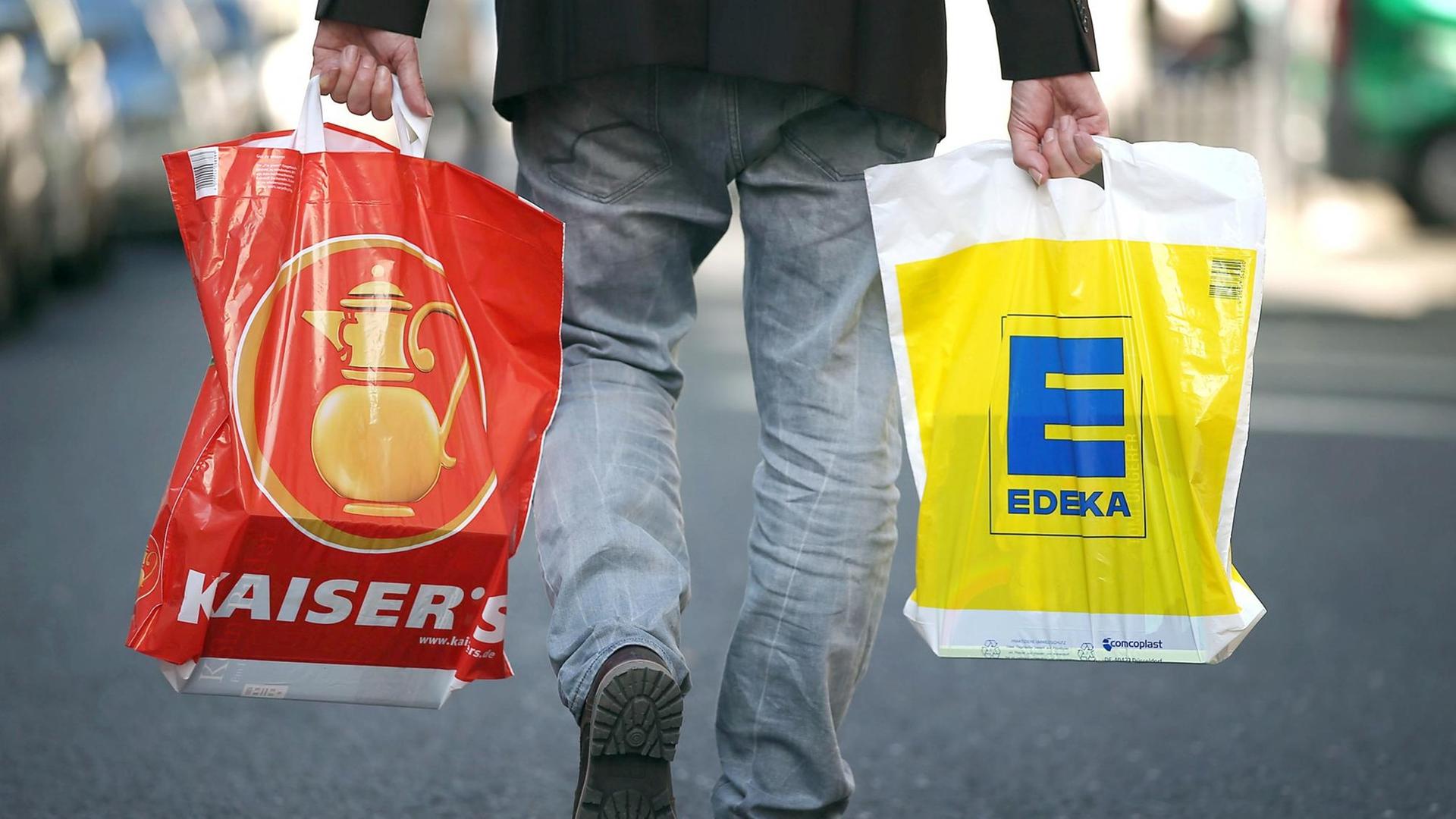 Ein Mann geht mit jeweils einer Tragetasche der Supermarktketten Kaisers's Tengelmann und Edeka über die Straße. Ein Mann geht mit jeweils einer Tragetasche der Supermarktketten Kaisers's Tengelmann und Edeka über die Straße.