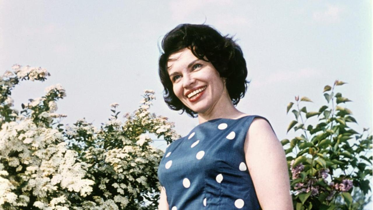 Inge Brandenburg steht in einem blauen Kleid mit großen weißen Punkten in einem Garten mit blühenen Büschen.