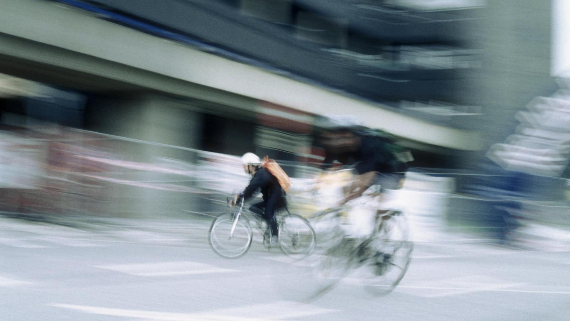 Zwei Fahrradkuriere sind vor einer Stadtkulisse zu sehen. Verwischte Elemente des Bildes vermitteln den Eindruck von Geschwindigkeit. Zwei Fahrradkuriere sind vor einer Stadtkulisse zu sehen. Verwischte Elemente des Bildes vermitteln den Eindruck von Geschwindigkeit.