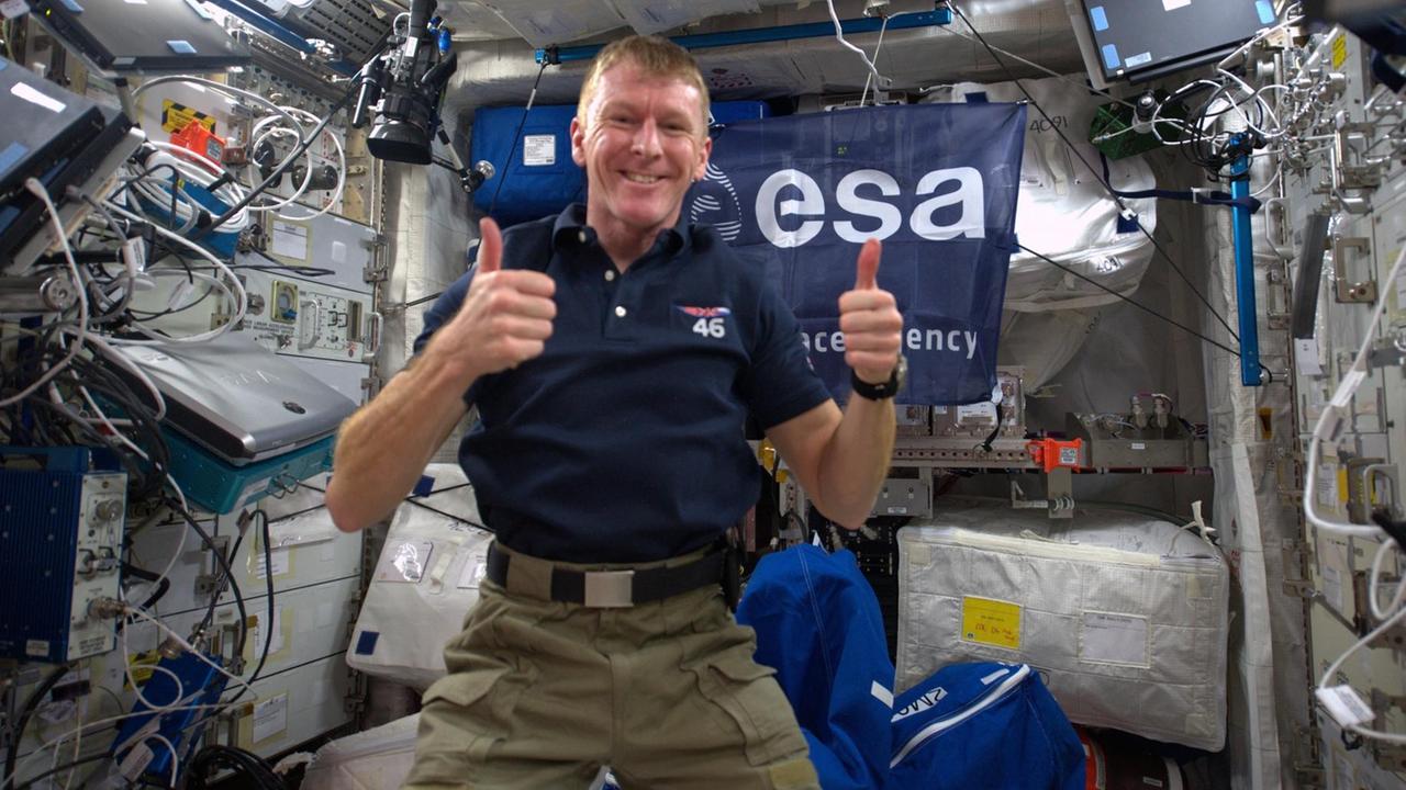 Peake schwebt im blauen T-Shirt in einem Raum voller elektronischer Geräte und Kabel und grinst mit erhobenen Daumen in die Kamera. Peake schwebt im blauen T-Shirt in einem Raum voller elektronischer Geräte und Kabel und grinst mit erhobenen Daumen in die Kamera.