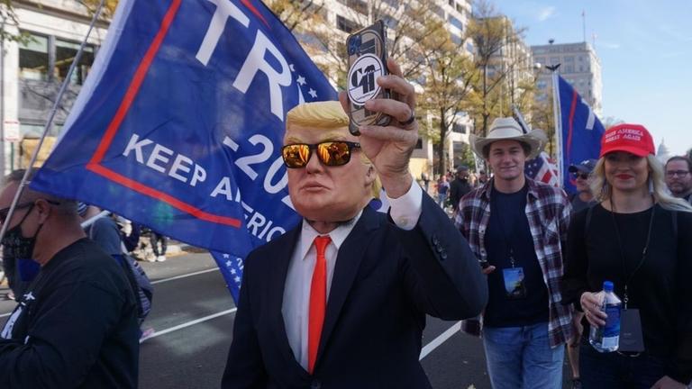 Ein Anhänger von Donald Trump trägt eine Maske Trumps und hält ein Smartphone in die Höhe Ein Anhänger von Donald Trump trägt eine Maske Trumps und hält ein Smartphone in die Höhe