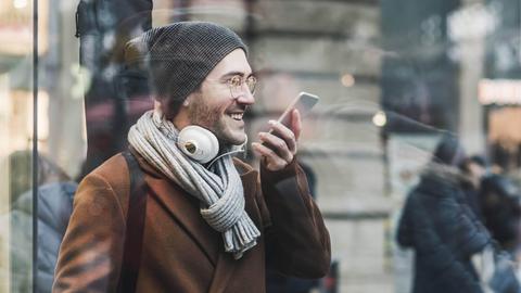 Ein lachender junger Mann mit Schal, Mütze und Gitarre auf dem Rücken spricht eine Nachricht in sein Smartphone (Symbolfoto)