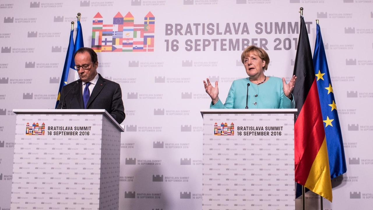 Merkel und Hollande stehen hinter Podesten vor einer helllilafarbenen Wand mit dem Gipfel-Logo. Merkel spricht und gestikuliert mit den Händen; Hollande hört zu.