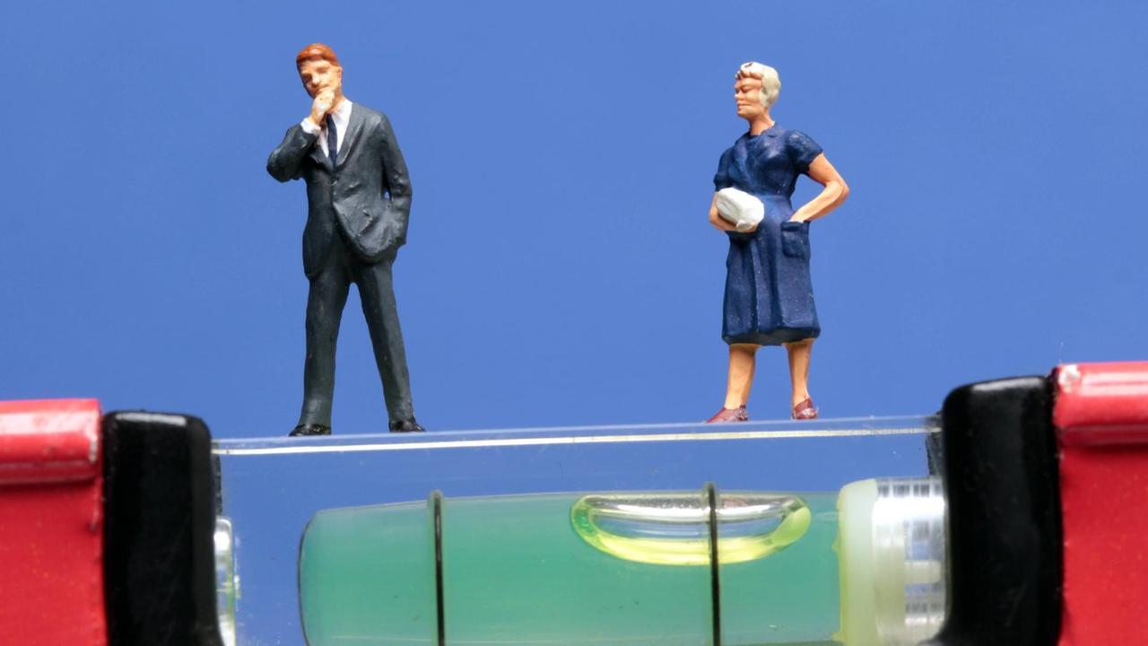 Zwei Miniatur-Figuren, eine männlich, eine weiblich, stehen auf einer schiefen Ebene einer Wasserwaage. Die männliche Figur steht etwas weiter unten. Zwei Miniatur-Figuren, eine männlich, eine weiblich, stehen auf einer schiefen Ebene einer Wasserwaage. Die männliche Figur steht etwas weiter unten.