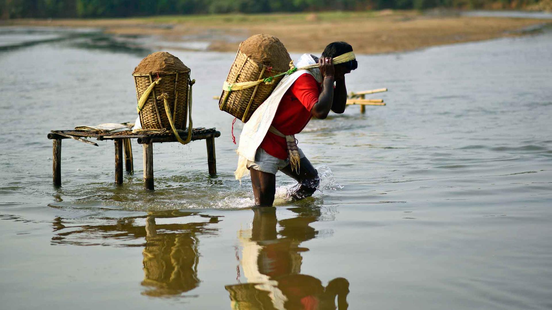 Eine Mann watet aus dem Wasser, auf dem Rücken trägt er einen Eimer voll Sand. Eine Mann watet aus dem Wasser, auf dem Rücken trägt er einen Eimer voll Sand.