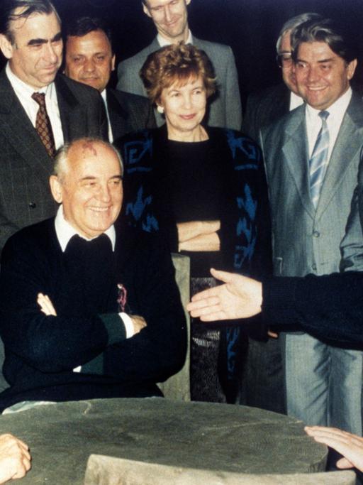 Bundeskanzler Helmut Kohl (r), der sowjetische Staatspräsident Michail Gorbatschow (M) und Bundesaußenminister Hans-Dietrich Genscher (l) unterhalten sich am 15.07.1990 in entspannter Atmosphäre an einem rustikalen Arbeitstisch in der freien russischen Natur, während die anderen Gäste unter anderem Raissa Gorbatschowa (M hinten) und Eduard Schewardnadse amüsiert die Szene betrachten.