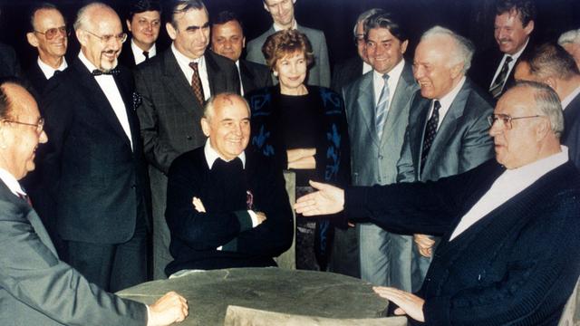 Bundeskanzler Helmut Kohl (r), der sowjetische Staatspräsident Michail Gorbatschow (M) und Bundesaußenminister Hans-Dietrich Genscher (l) unterhalten sich am 15.07.1990 in entspannter Atmosphäre an einem rustikalen Arbeitstisch in der freien russischen Natur, während die anderen Gäste unter anderem Raissa Gorbatschowa (M hinten) und Eduard Schewardnadse amüsiert die Szene betrachten.