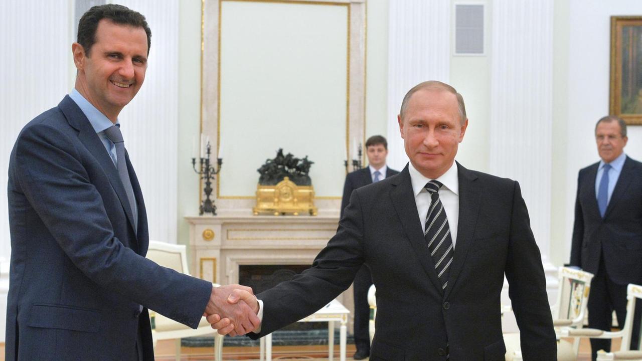 Assad und Putin schütteln sich im weißen Empfangsraum des Kreml die Hände und lächeln in die Kamera. Im Hintergrund stehen der russische Außenminister Lawrow und eine weitere Person. 