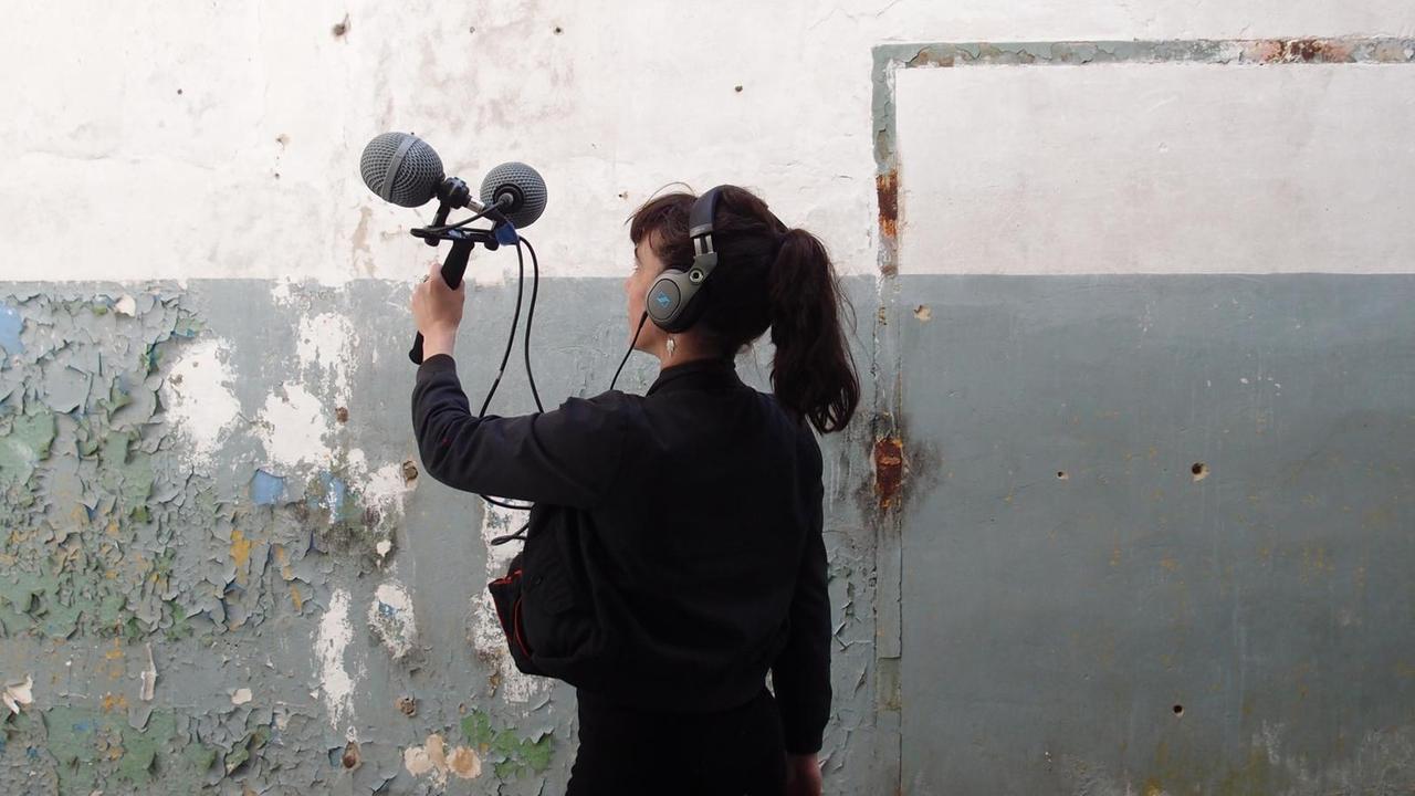Die Klangkünstlerin Marie Guérin steht vor einer Wand im Halbmondlager in Wünsdorf und nimmt mit einem Mikrofon auf.