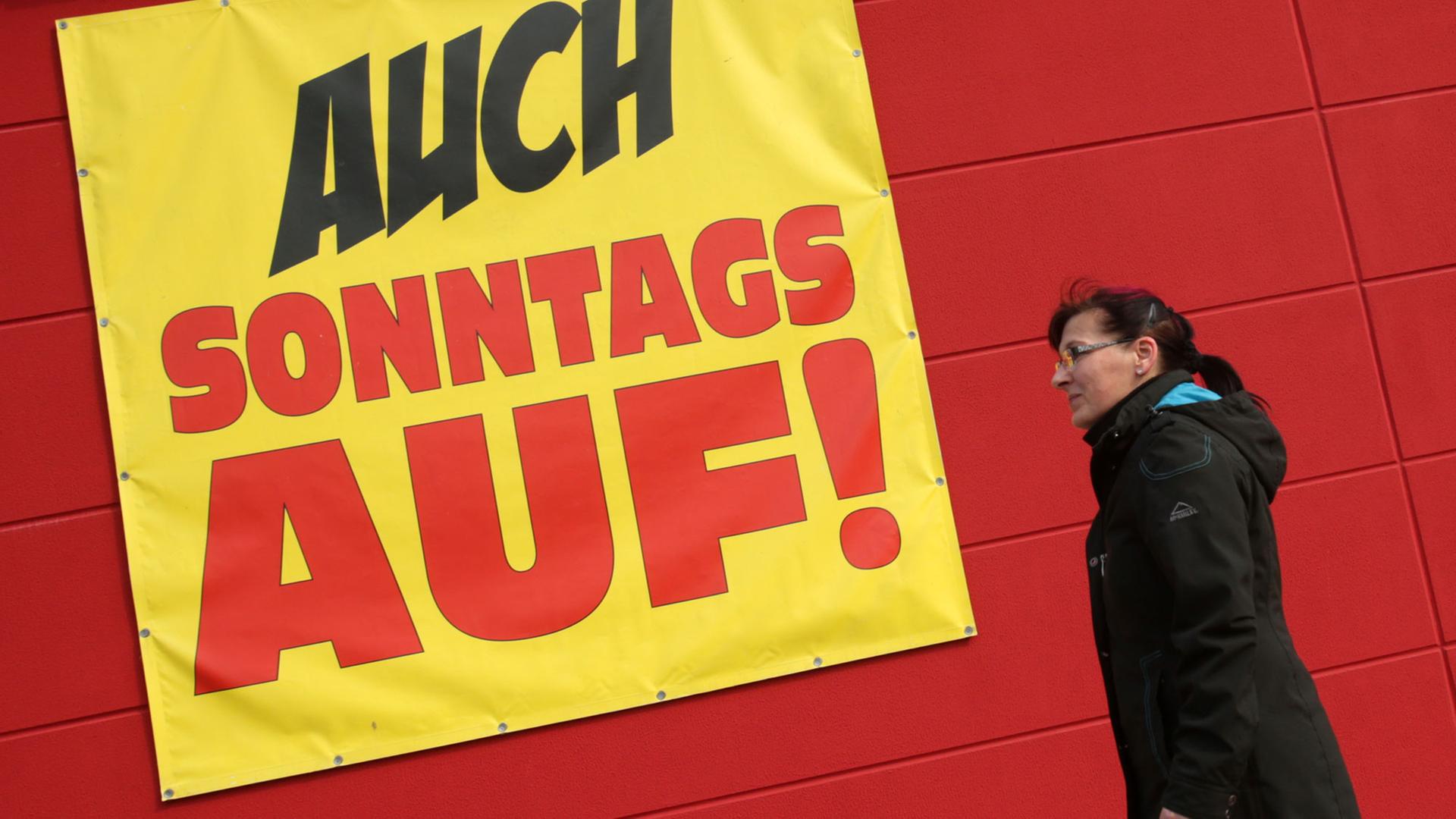 Eine Frau mit einer Einkaufstüte in der Hand geht an einem Schild vorbei, auf dem steht "Auch sonntags offen". Eine Frau mit einer Einkaufstüte in der Hand geht an einem Schild vorbei, auf dem steht "Auch sonntags offen".