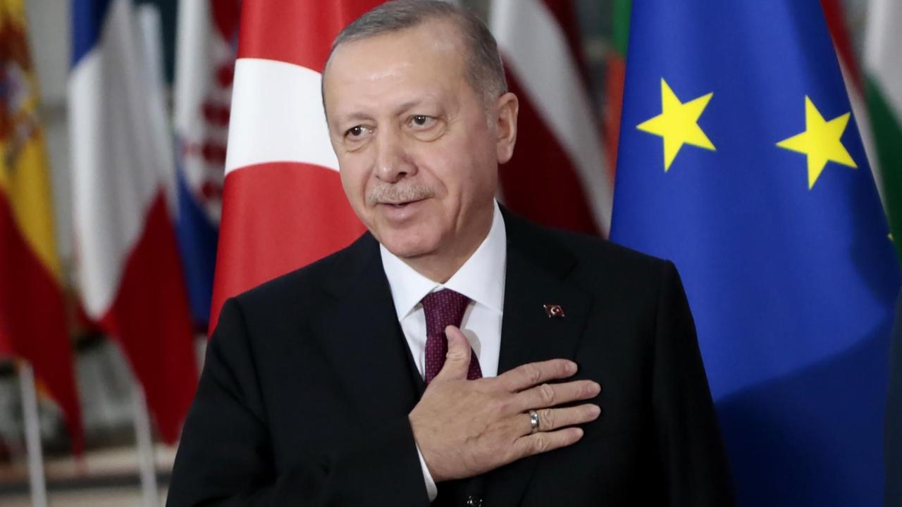 Erdogan in Jacket und mit Krawatte legt die Hand aufs Herz und steht vor der Fahne der EU.
