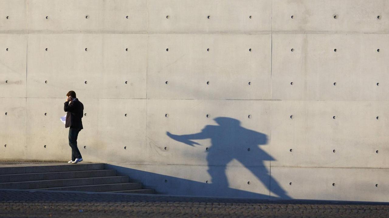 Eine Mann im Anzug telefoniert vor einer Betonwand und wirft dabei einen Schatten.
